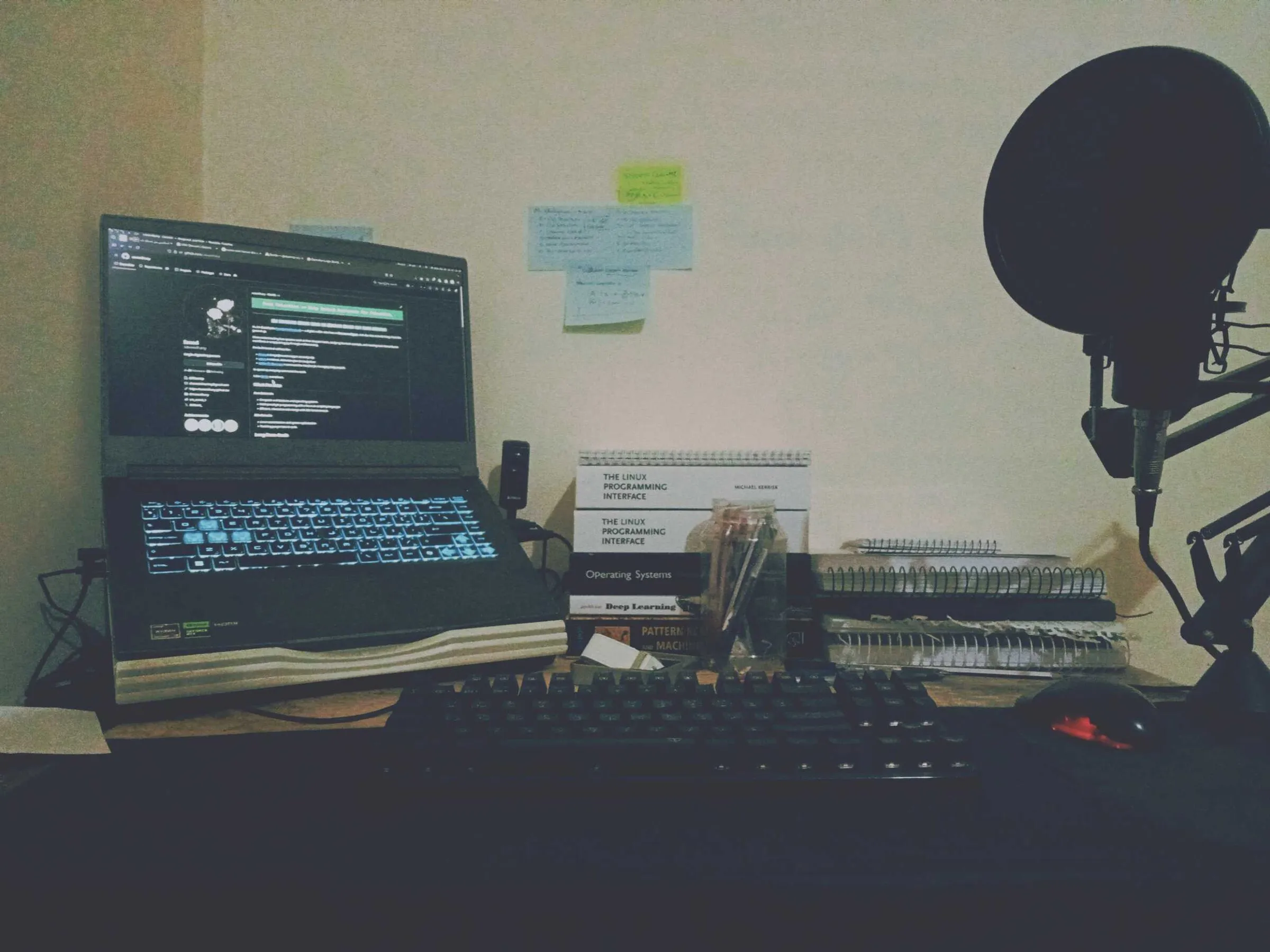 A laptop, a microphone on a boom arm and a stack of programming books on a desk against a pale wall